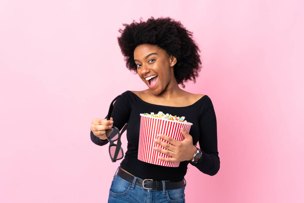 Young African American woman isolated on pink background with 3d glasses and holding a big bucket of popcorns - Fotografie, Obrázek