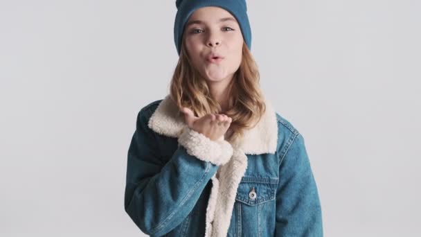 Bella ragazza bionda adolescente vestita con giacca e cappello in denim che invia baci d'aria in camera isolata su sfondo bianco. Affascinante adolescente in posa in studio - Filmati, video