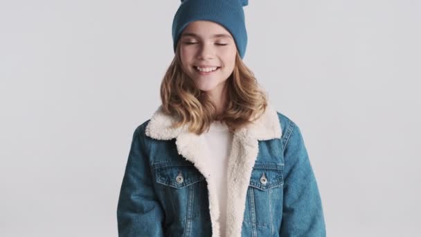 Csinos szőke tinédzser lány farmerdzsekiben és kalapban, igent integetve a kamerának, fehér háttér felett mosolyogva. Egyetértés - Felvétel, videó
