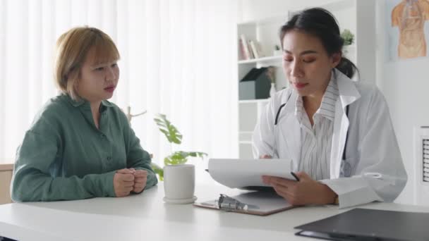 Jonge Asia vrouwelijke arts in wit medisch uniform met behulp van klembord is het leveren van grote nieuws talk bespreken resultaten of symptomen met meisje patiënt zit aan de balie in de kliniek of ziekenhuis kantoor. - Video