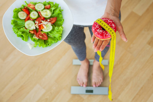 Διατροφή έννοια, η γυναίκα μετρά το βάρος σε ηλεκτρονική κλίμακα, ενώ κρατώντας θερμίδες ντόνατ - Φωτογραφία, εικόνα