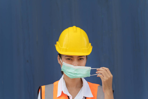 Trabajadora asiática se para en la pared azul. Trabajadora de trabajo está usando la máscara de protección de la cara y el casco de seguridad y el uso de naranja reflectante vestido de seguridad  - Foto, Imagen