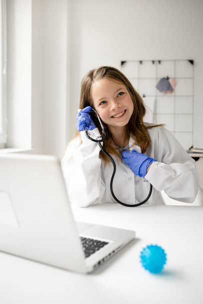 pretty blonde and young schoolgirl is playing doctor and has white coat on and blue protective gloves and use a stethoscope - Foto, Imagen