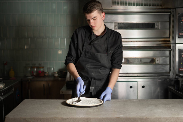 Szef kuchni w rękawiczkach przygotowuje pizzę w zakładzie produkcyjnym. Za nim są piekarniki elektryczne. Sprzęt i proces gotowania. - Zdjęcie, obraz