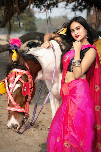 Retrato al aire libre de una chica india muy hermosa con saree y sostiene el caballo ensillado de riendas y posando de moda en un fondo borroso. Estilo de vida y moda. - Foto, imagen