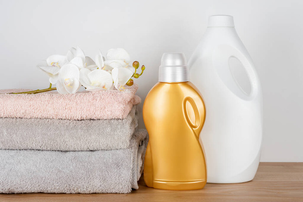 Bottiglie di detergente e ammorbidente per tessuti con asciugamani puliti e fiori di orchidea sul tavolo di legno. Contenitori di prodotti per la pulizia. Detergente liquido e condizionante. Giorno di lavanderia, concetto di pulizia - Foto, immagini