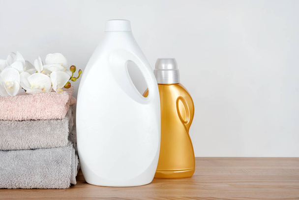 Butelki detergentu i zmiękczacza tkaniny z czystymi ręcznikami i kwiatami storczyków na drewnianym stole. Pojemniki środków czyszczących. Płynny detergent i odżywka. Dzień pralni, koncepcja czyszczenia - Zdjęcie, obraz