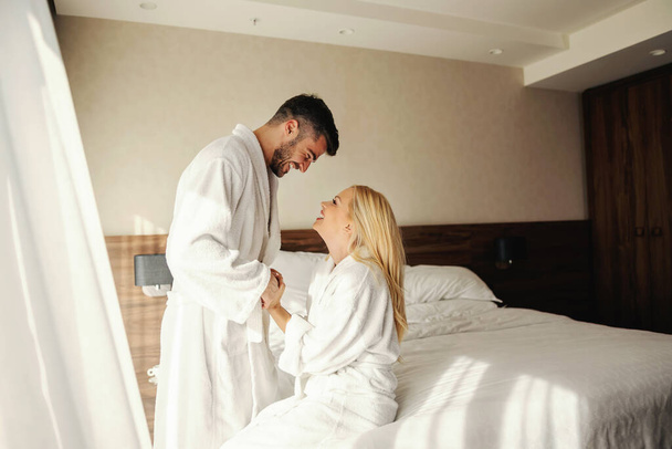 Waking up with a smile, mornings with a loved one. Sunny mornings in a room of a modern hotel, close couple in white bathrobes share a moment to remember. He holds her hands and looks her in the eye - Photo, Image