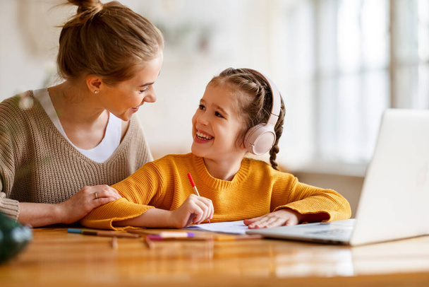 Vidám kis iskolás lány a fejhallgató írás gyakorlatok a copybook, miközben tanul távolról laptopon keresztül otthon boldog anya ül a közelben, és támogatást nyújt - Fotó, kép