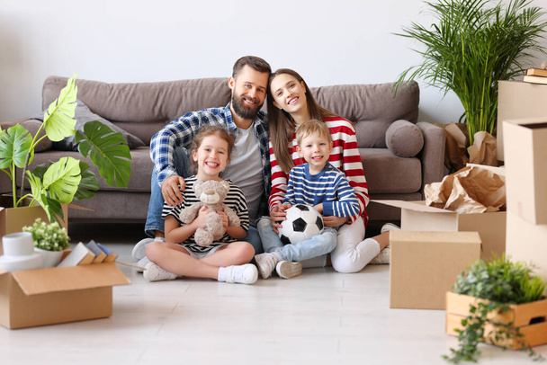 Delighted parents and cute kids gathering on floor in living room with unpacked boxes in new flat and looking at camera - Fotoğraf, Görsel