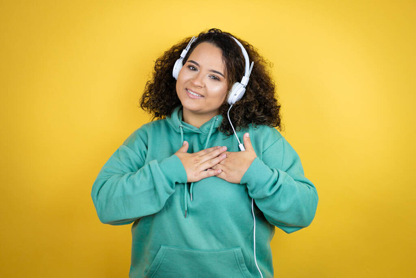 Fiatal afro-amerikai lány tornaruhát visel és fejhallgatót használ, mosolygós kezekkel a mellkasán és hálás gesztussal az arcán.. - Fotó, kép