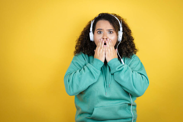 Jong Afrikaans-Amerikaans meisje draagt sportkleding en gebruikt een koptelefoon geschokt over de mond met handen voor een fout. geheim concept. - Foto, afbeelding