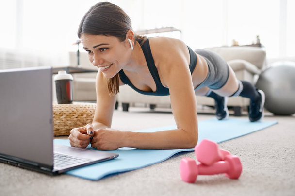 Woman doing abs exercise, online fit training at the laptop. Female person in sportswear, internet sport workout, room interior on background - Zdjęcie, obraz