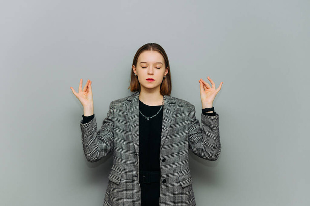Mujer joven atractiva en una chaqueta y ropa casual inteligente medita con las manos levantadas y los ojos cerrados sobre un fondo gris. Trabajadora de oficina se calma con yoga - Foto, imagen