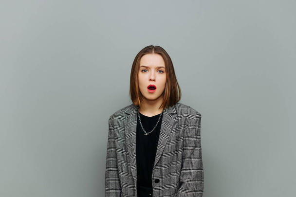 Portret van geschokte jonge zakenvrouw in slimme casual kleding, het dragen van jas, kijken naar camera met verrast gezicht op grijze achtergrond. - Foto, afbeelding