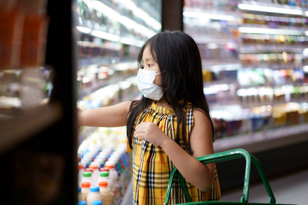 アジアの子供や子供の女の子は、スーパーマーケットやデパートで牛乳を購入するためのバスケットを保持してコロナウイルスcovid-19やほこりを保護するために病気に近い口の鼻やインフルエンザ咳のための白い顔のマスクを身に着けています - 写真・画像