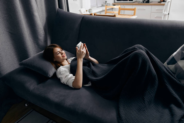 Eine Frau mit Handy liegt auf einem grauen Sofa neben dem Fenster im Hintergrund - Foto, Bild