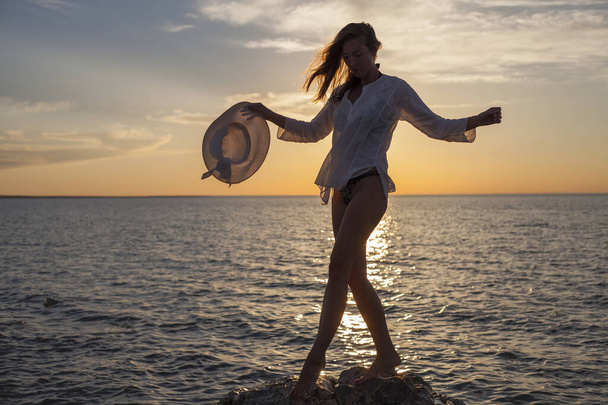 Силуэт молодой счастливой беззаботной женщины, позирующей в шляпе на пляже на закате - Фото, изображение