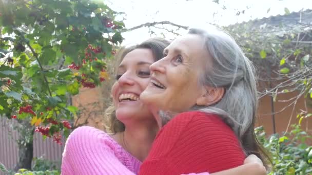 Vanhempi äiti harmailla hiuksilla aikuisen tyttärensä kanssa katsomalla kameraa puutarhassa ja halaamalla toisiaan aurinkoisena päivänä ulkona - Materiaali, video