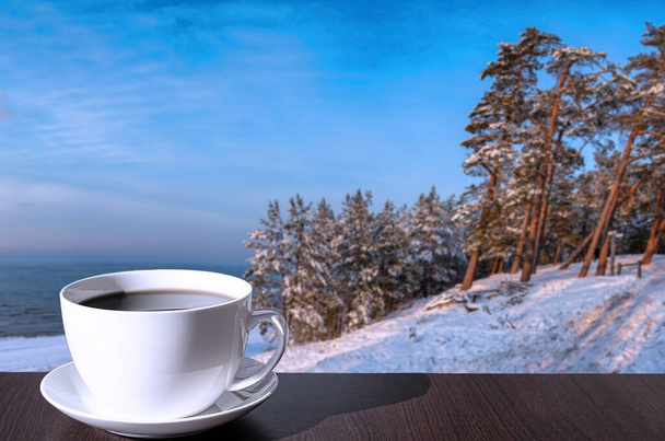 Cup of coffee on the table with view of snowy beach, Baltic sea and covered in snow fir and pine trees on the hill. - Photo, Image