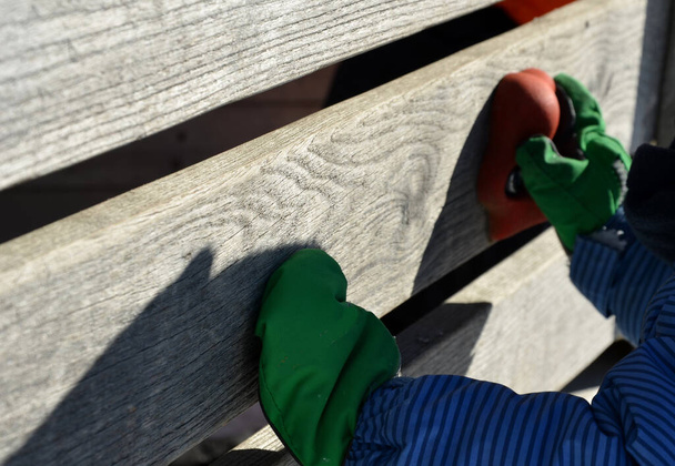 μικρό παιδάκι σκαρφαλώνει σε έναν τεχνητό τοίχο αναρρίχησης με πολύχρωμες λαβές. Μικρό αγόρι χέρι κατέχουν πολύχρωμες πέτρες σε ξύλινο πίνακα. μικρό αγόρι με μπουφάν χειμώνα και αθλητικά γάντια ηλιόλουστη μέρα - Φωτογραφία, εικόνα