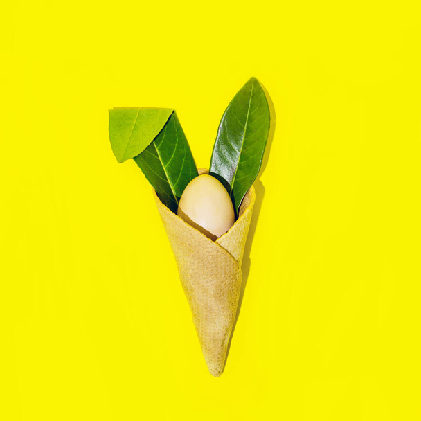 Lapin de Pâques dans un cône avec des oreilles de feuilles et un oeuf. Sur fond jaune, composition minimale. - Photo, image