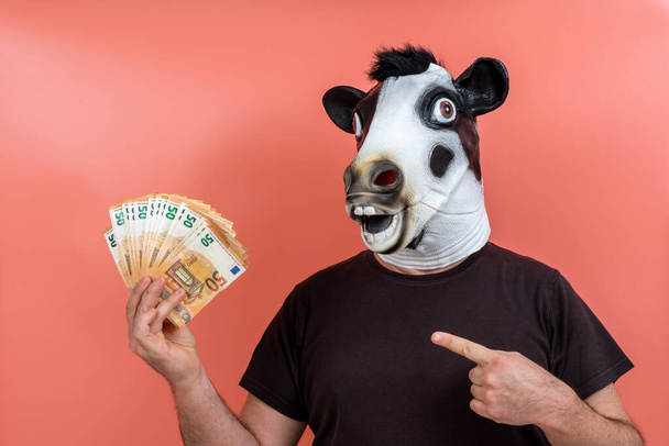 boy with funny latex cow mask holding money with one hand and pointing to it with the finger of the other hand and pink background - Photo, Image