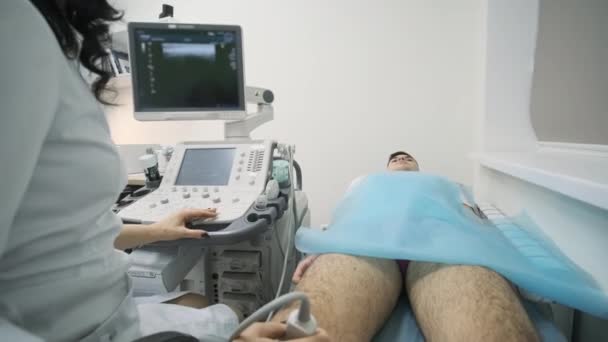 Kadın doktor elleri, genç adamın diz eklemlerini ultrason cihazı ve ultrason cihazı kullanarak inceliyor. Doktor, hastanın bacağına ve dizine ultrason sensörü uygular.. - Video, Çekim