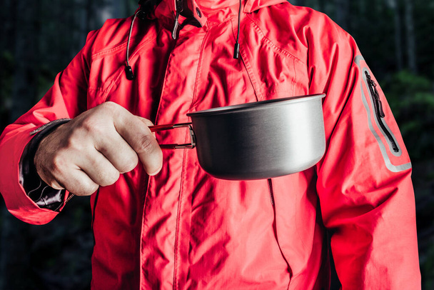 金属カップを保持する赤い旅行キャンプジャケットでハイカーの男のクローズアップ製品写真. - 写真・画像
