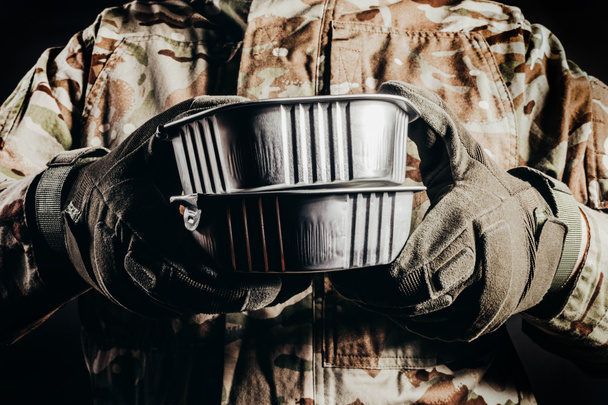 Foto van soldaat in gecamoufleerd uniform en tactische handschoenen met ingeblikt voedsel MRE op zwarte achtergrond, close-up zicht. - Foto, afbeelding