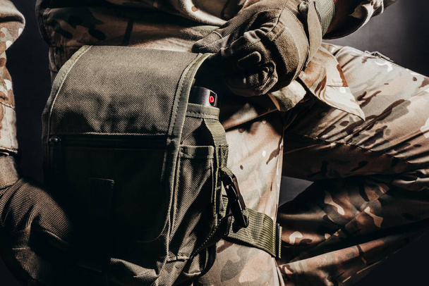 Foto des sitzenden Soldaten in getarnter Uniform und taktischen Handschuhen mit Beintasche auf schwarzem Hintergrund. - Foto, Bild