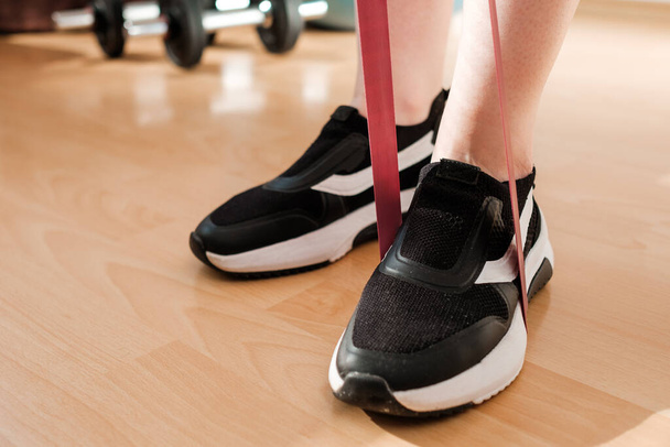 Женские ноги в спортивных леггинсах и кроссовках делают упражнения с фитнес-эластичными лентами дома во время изоляции. Домашняя тренировка. с полосами сопротивления.  - Фото, изображение
