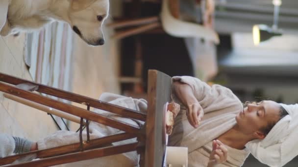 Verticale schot van jonge vrouw in badjas en handdoek wrap op hoofd zitten aan tafel in de woonkamer en het geven van stuk taart aan schattige golden retriever hond - Video
