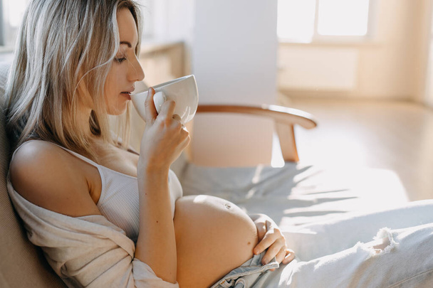 Pregnant woman sitting on a sofa at home, drinking a cup of coffee or tea. - Foto, Imagem