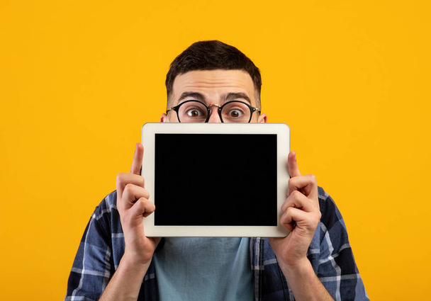 Shocked young guy hiding behind tablet computer with blank screen on orange studio background, mockup for design - Photo, Image