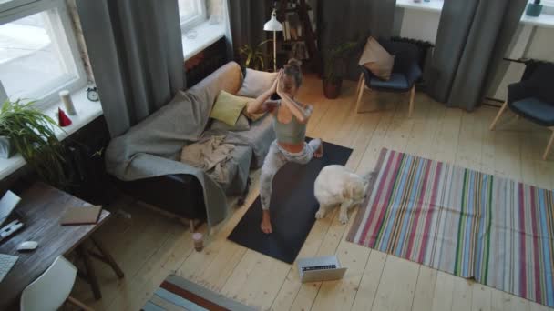 Savaşçı pozu veren zayıf bir kadının yüksek açılı çekimi ve evde köpekle antrenman yaparken bilgisayardan yoga dersi izlemesi. - Video, Çekim