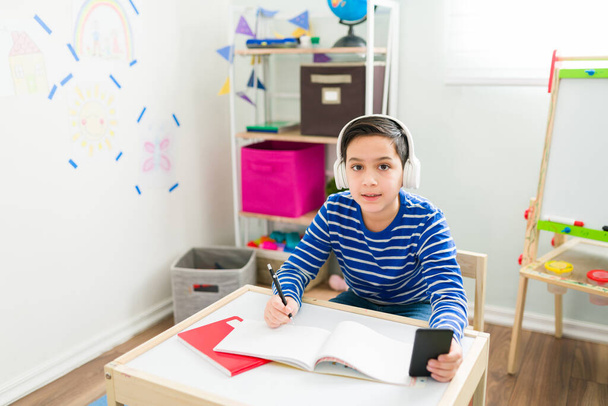 Biały chłopiec ze słuchawkami trzymający smartfon i piszący w notatniku siedząc na małym biurku. Happy elementarny chłopiec o wirtualnej klasy online - Zdjęcie, obraz