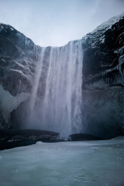 Vista ad angolo basso sulla cascata Skogafoss alta sessanta metri in Islanda. Acqua che si riversa su scogliere ghiacciate in un remoto paesaggio invernale nordico. - Foto, immagini