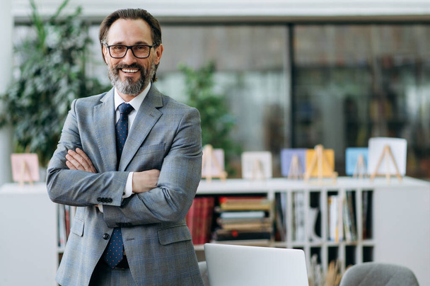 Portrait of a confident middle aged entrepreneur in stylish suit and eyeglasses standing in modern office, looking at the camera and friendly smiling - Photo, Image