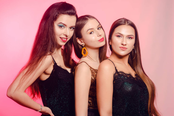 3 Mädchen posieren lachend auf rosa Hintergrund. Mädchen in brillanten Kleidern mit verschiedenfarbigem Make-up. Drei brünette Mädchen tanzen und genießen das Leben. 8. März, Frauenpower, Frauentag - Foto, Bild