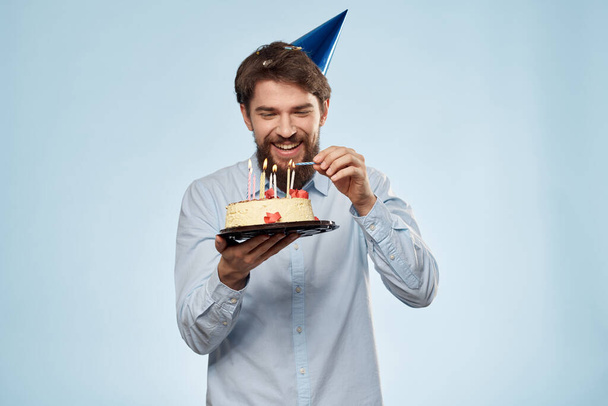 Uomo barbuto con un piatto di torta su sfondo blu e un cappello da festa di compleanno sulla testa - Foto, immagini