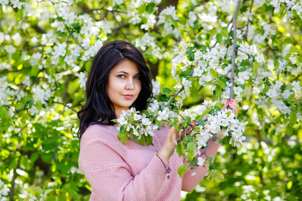 Portrait of a fat plump young woman with black hair and a blooming apple tree on a white floral background in a park on a spring day. Happy girl plus size. - Photo, Image
