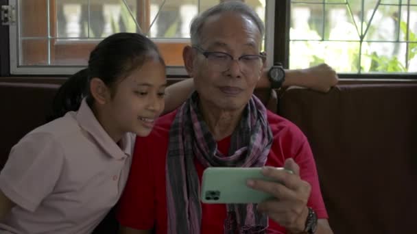 Schattige kleindochter nadert en zit naast haar oudere grootvader en kijkt online sociale media vanaf een mobiele smartphone. Samenzijn in een gezin van twee generaties. - Video