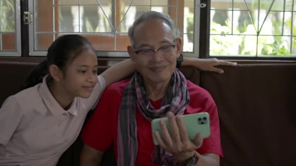 Rodzina w dwóch pokoleniach ze starszym dziadkiem i uroczą wnuczką dołączających i oglądających filmy społecznościowe online ze smartfona komórkowego w salonie.  - Materiał filmowy, wideo