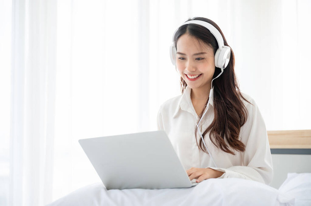 Feliz bela jovem mulher asiática trabalhando no computador portátil e sentado na cama em casa. Ela fazendo webinar videoconferência e usando fone de ouvido - Foto, Imagem