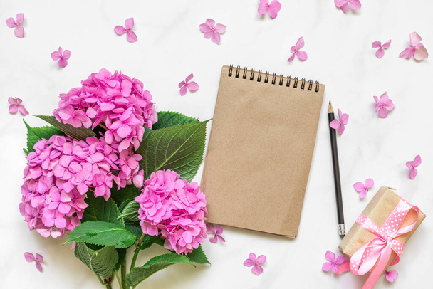 Blanco papieren notitieboekje in bloemblaadjes frame met roze hortensia bloemen boeket en geschenkdoos op witte achtergrond. Maak je klaar. Vlakke wip. Bovenaanzicht. bruiloft of vakantie concept. feestelijke achtergrond - Foto, afbeelding