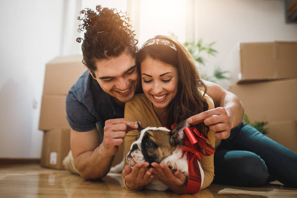 Ein glückliches junges Paar macht eine Pause und hat Spaß mit seinem englischen Bulldoggen-Welpen beim Einzug in sein neues Zuhause. - Foto, Bild