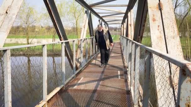 Fogyatékos szakállas öregember sétapálcával lassan mozog, és figyeli a fém hídon a kis folyó felett - Felvétel, videó