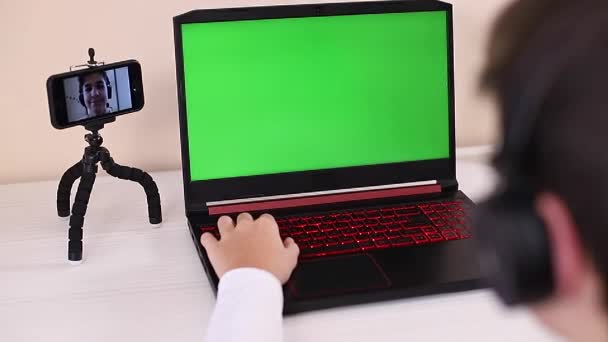 Vue arrière du jeune garçon jouer jeu sur ordinateur portable. Écran vert dans l'ordinateur portable. Steamer dans les écouteurs capturer des vidéos sur son téléphone sur le trépied. Maquette-toi. Ordinateur portable en bref - Séquence, vidéo