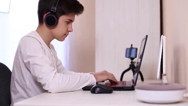 Мальчик-подросток играет в игру на компьютере в белой комнате. Снимать видео на веб-камеру на ноутбуке - Кадры, видео